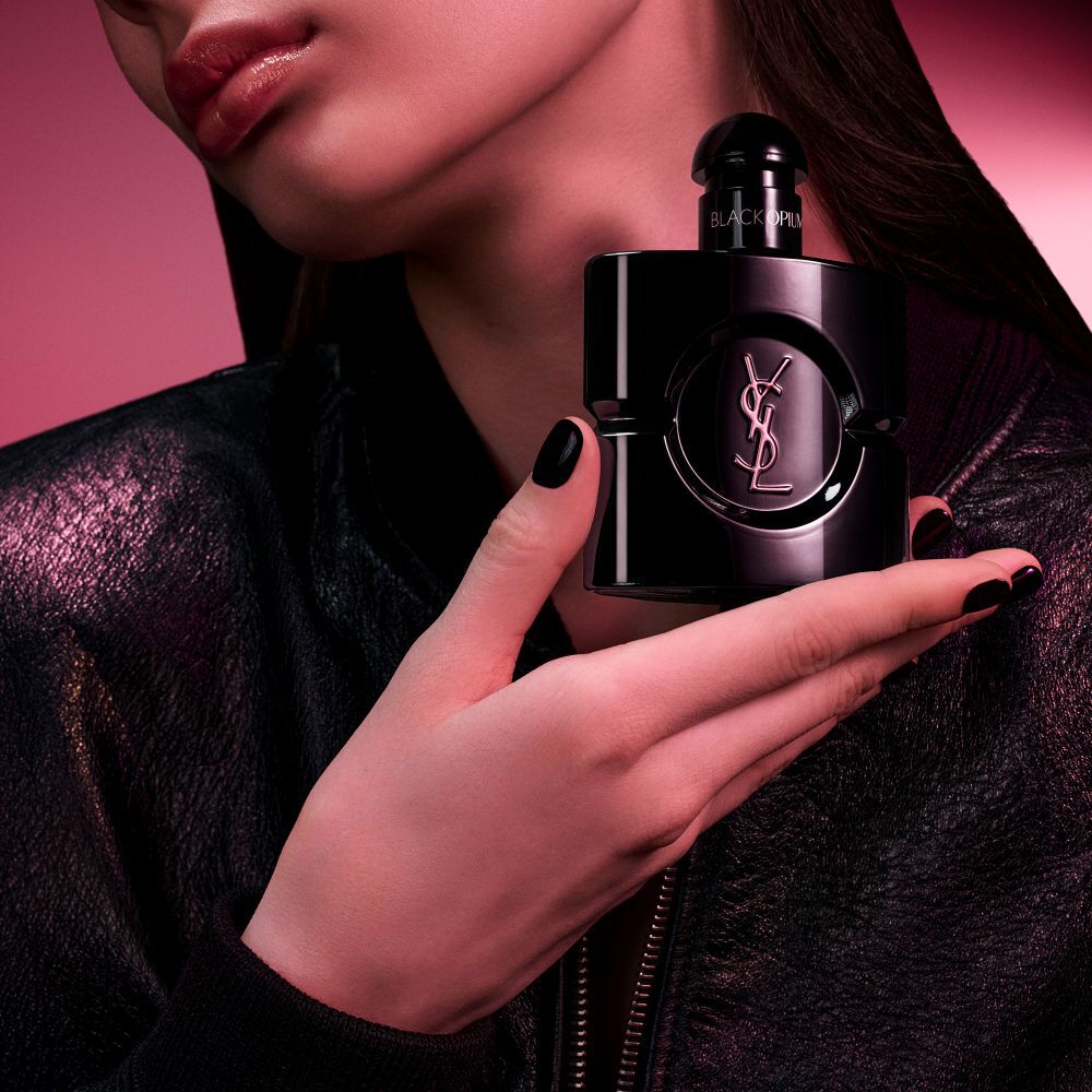 Black Opium Le Parfum Flakon in glanzendem Schwarz, umgeben von dunklen Vanilleschoten und weißem Jasmin mit Produktversprechen.