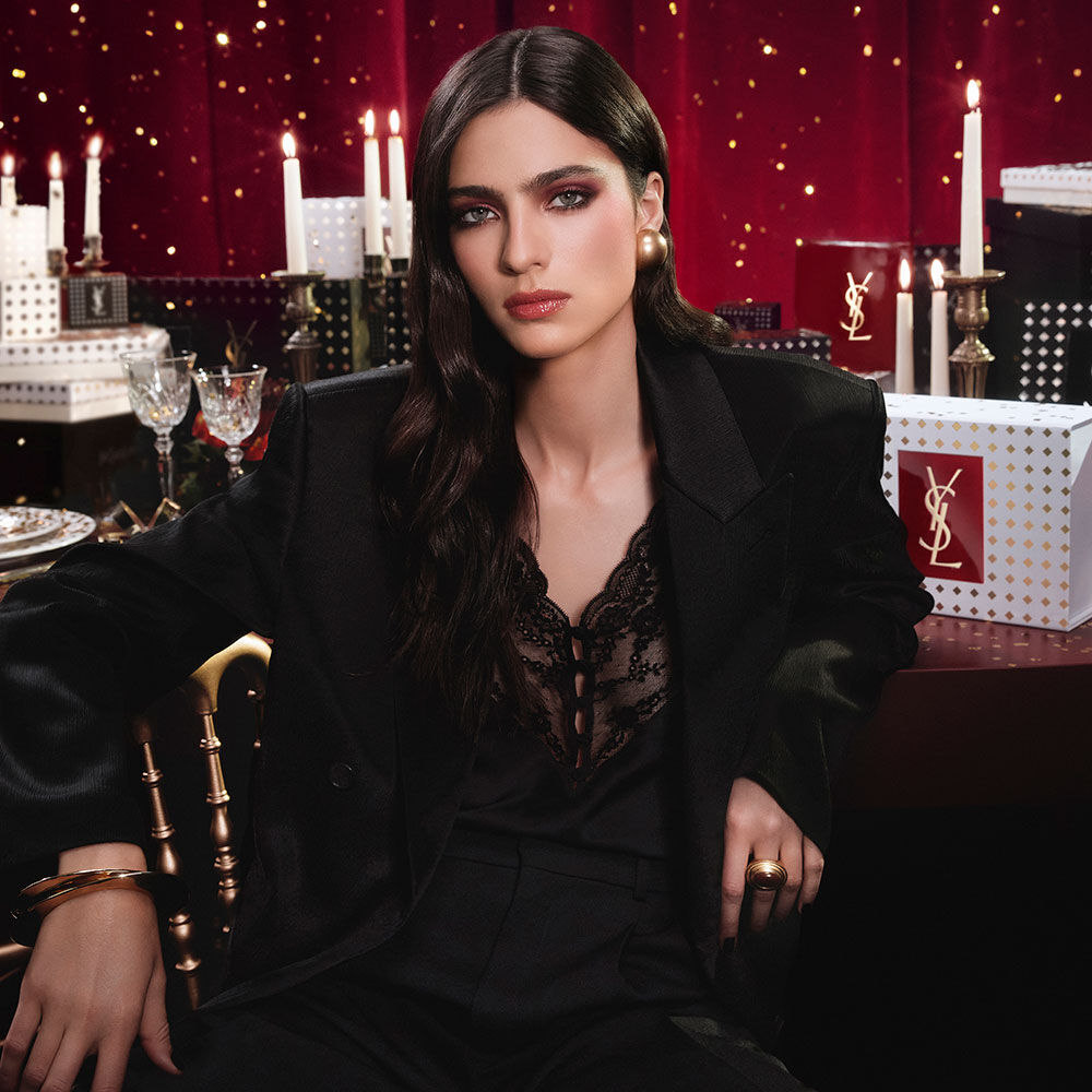 Modell in schwarzem Anzug und glamourosem Look, sitzend in festlichem Kerzenlicht mit YSL-Geschenkboxen.