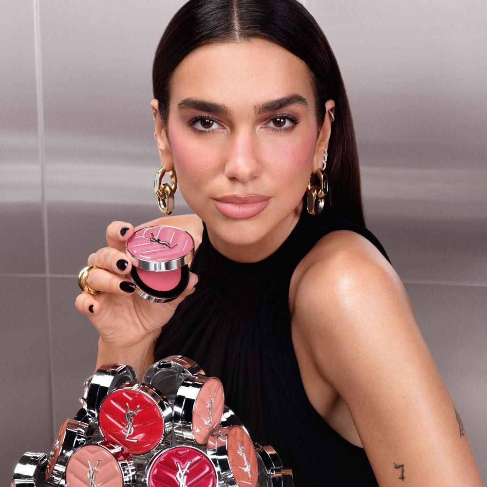 Dua Lipa halt ein geoffnetes YSL Make Me Blush Lifted Glow Cream Blush Kompakt in der Hand – vor ihr eine Kollektion der neuen Nuancen.