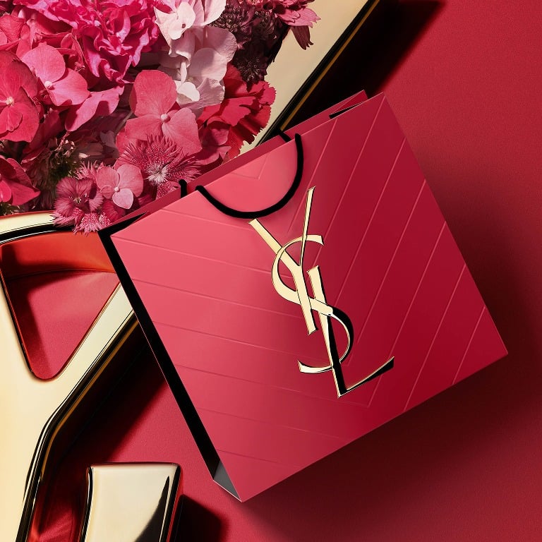 YSL-Geschenkbox in leuchtendem Rosa mit goldenem Cassandre-Logo, umgeben von frühlingshaften Blütenblättern.