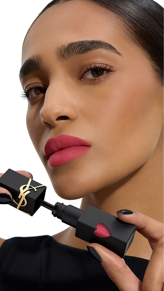 Modell mit mittlerem Hautton und einer intensiven Rosen-Matt-Lippenfarbe, die die schwarze Flasche von YSL THE INKS BLUR Lip Stain mit ihrem pinken Herz hält.