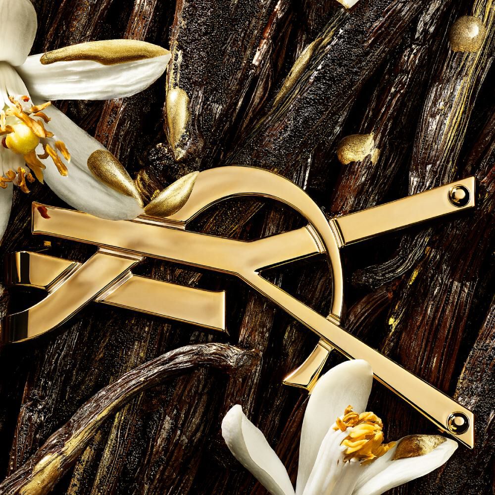 Nahaufnahme des goldenen YSL-Logos, platziert zwischen Vanilleschoten, weißen Blüten und funkelnden goldenen Akzenten.