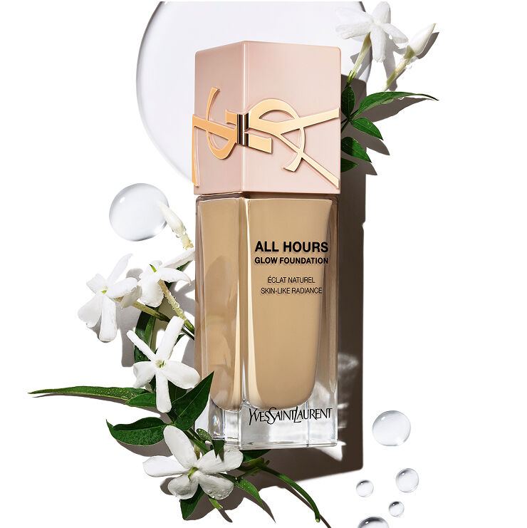 YSL All Hours Glow Foundation Flasche mit 'Éclat Naturel Skin-Like Radiance' umgeben von weißen Jasminblüten und klaren Wassertropfen, die natürliche Inhaltsstoffe und Hydratation suggerieren.