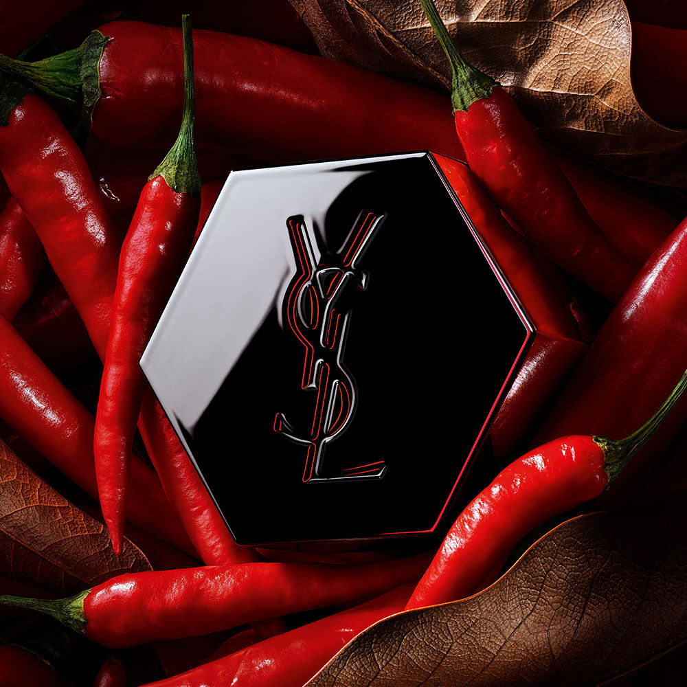 Schwarzer sechseckiger YSL-Parfümverschluss mit rot umrandetem Logo, umgeben von roten Chilischoten.