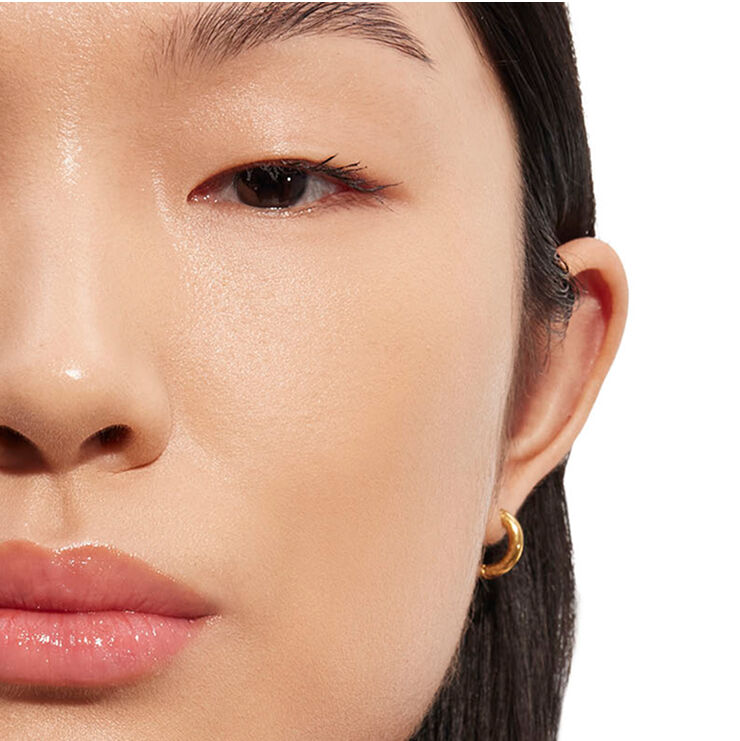 Detaillierte Nahaufnahme des Gesichts einer Person, mit Fokus auf Augen- und Wangenbereich mit natürlich aussehendem Make-up und einem goldenen Creolenohrring, der die Hautleuchtkraft demonstriert.