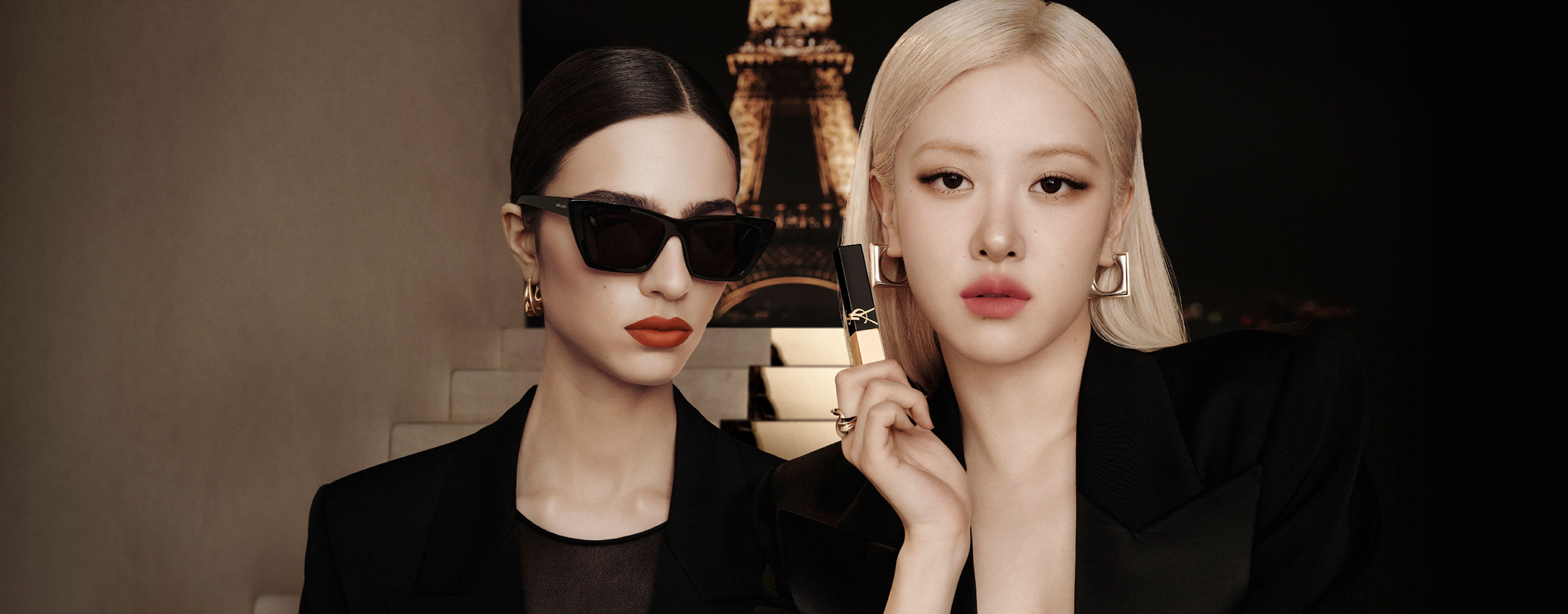 Zwei elegant in Schwarz gekleidete Models vor nächtlicher Pariser Kulisse mit Eiffelturm; eine hält einen schwarz-goldenen YSL-Lippenstift, beide mit markanten Ohrringen und glatten Frisuren.