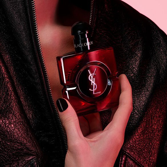 Nahaufnahme einer Hand mit schwarzen Nägeln, die den Flakon Black Opium Eau de Parfum Over Red vor einer schwarzen Lederjacke hält.