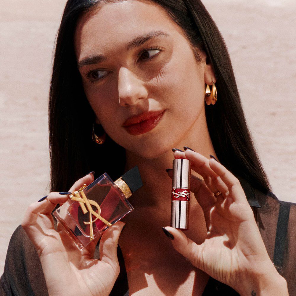 Dua Lipa hält Libre Eau de Parfum Berry Crush und einen YSL Loveshine, mit roten Lippen und goldenen Ohrringen.